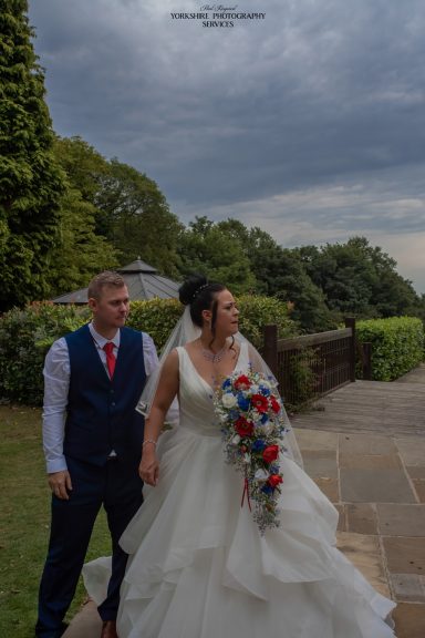 Bride and groom holding hands, smiling outdoors with cloudy skies and greenery in the background.