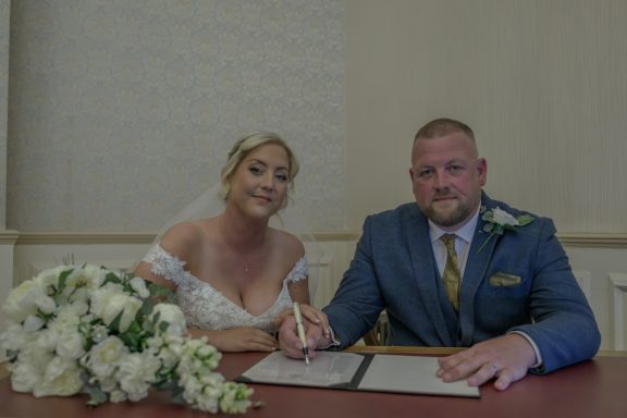 Bride and groom signing a marriage register with a floral bouquet nearby.