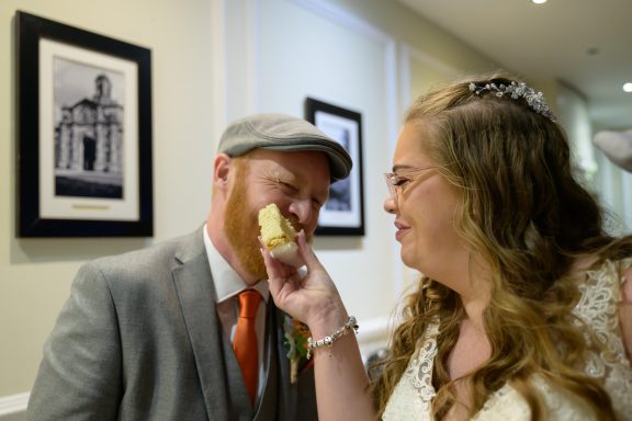 Bride playfully feeding cake to groom, both smiling, in a decorated venue.