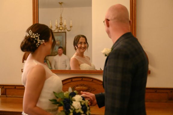 Bride smiling at her reflection in a mirror while standing with the groom.