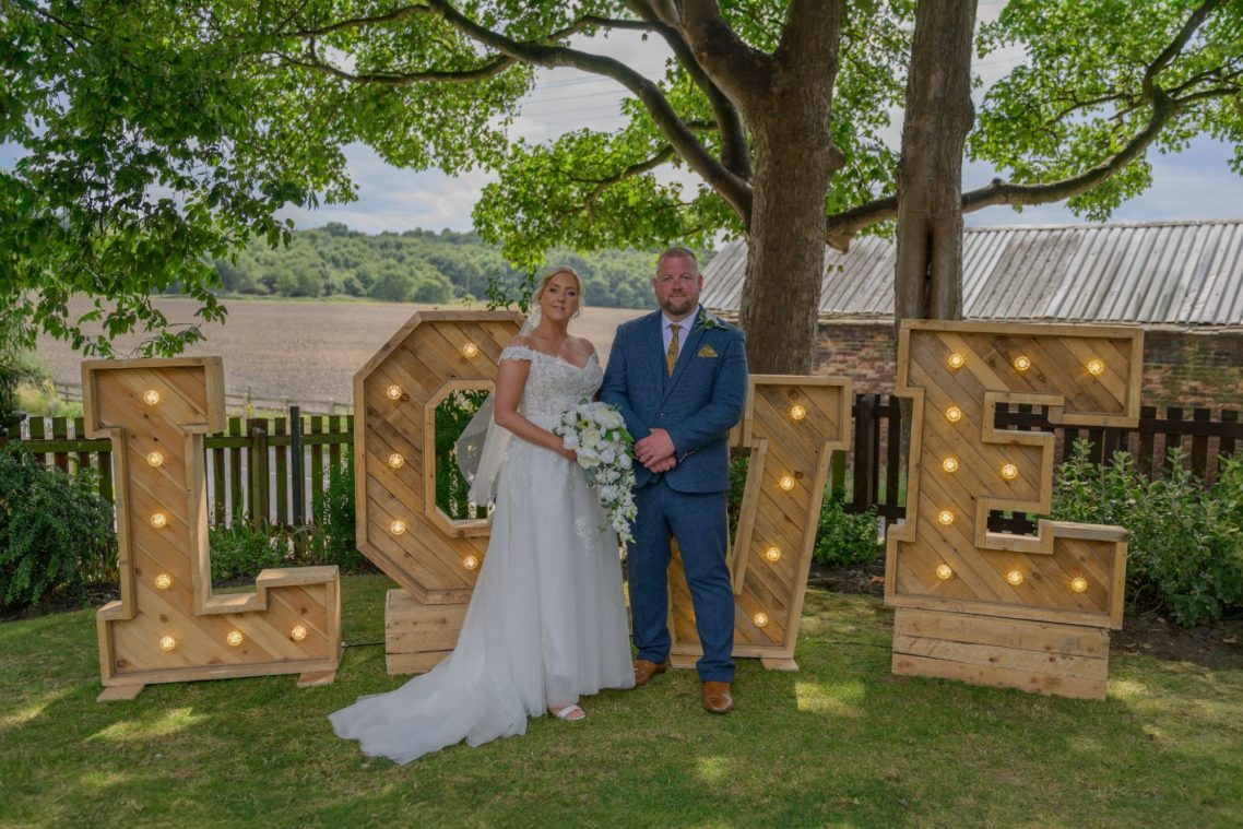 Bride and groom stand beside large illuminated letters spelling "LOVE" in a sunny garden setting.