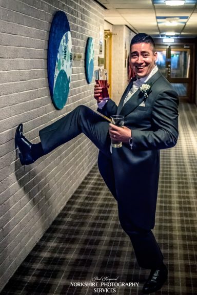 A smiling man in a suit poses playfully with one leg raised, holding a drink.